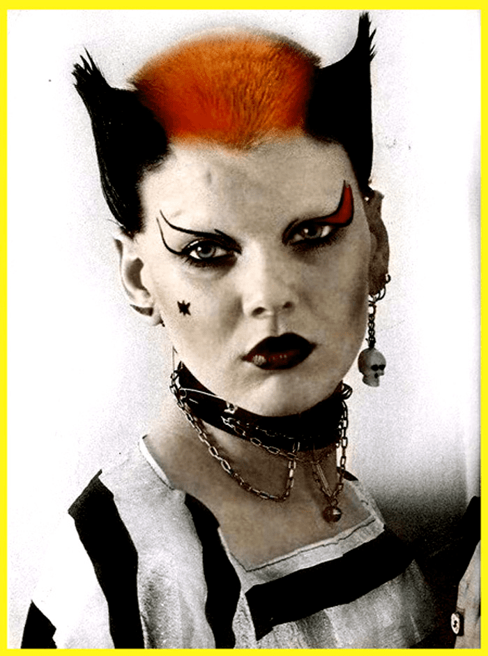 Soo Catwoman is serious while standing, has spiky black hair and orange in the middle, funky makeup, a star on her right cheek, wearing skull earrings on her left ear and horn earing on right with a gold chain with a bell on a black choker, black and white striped shirt.