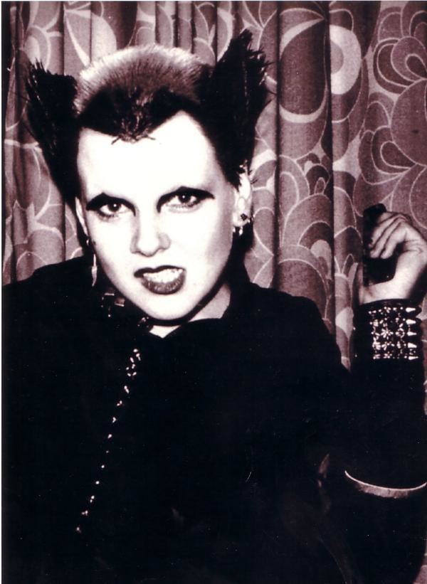 Soo Catwoman is serious, mouth half open showing its teeth while holding a comb on her left hand. has spiky black hair and funky makeup, a dot on her right cheek wearing black and white earrings, a spiky punk bracelet, ring bracelet, and black jacket.
