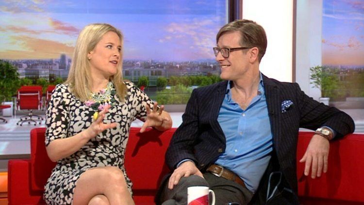 Sophie Robinson and Daniel Hopwood are smiling while talking to each other and sitting on a red couch with crossed legs in the set of "The Great Interior Design Challenge". Sophie with blonde hair, wearing a necklace and a black and white dress while Daniel is wearing eyeglasses, a wrist watch on his left hand, a black coat with a blue handkerchief over a blue long sleeves, a brown belt, and gray pants.