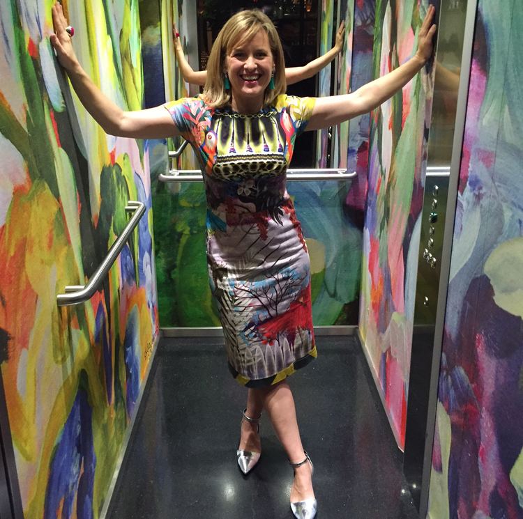 Sophie Robinson is smiling while her hands are on the colorful wall of an elevator, with blonde hair, wearing earrings, a multi-colored dress, and silver sandals.