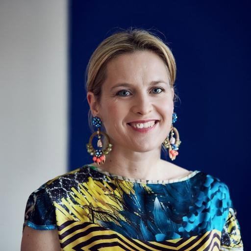 Sophie Robinson is smiling, with blonde hair, wearing a colorful chandelier style earrings and a multi-colored dress.