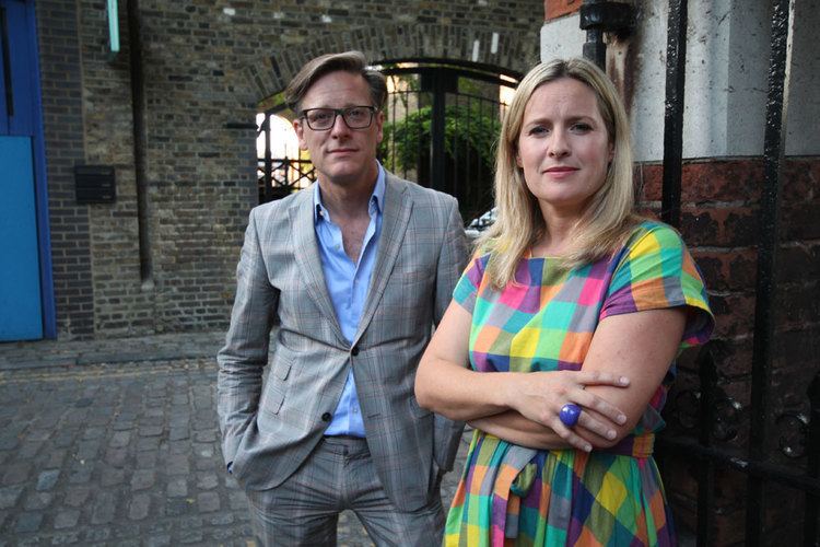 Sophie Robinson with a tight-lipped smile with hands crossed on her chest while Daniel Hopwood with a serious face with hands in his pocket in the set of "The Great Interior Design Challenge". Sophie with blonde hair, wearing a blue ring and a multi-colored dress while Daniel is wearing eyeglasses, a gray checkered coat over blue long sleeves, and gray checkered pants.