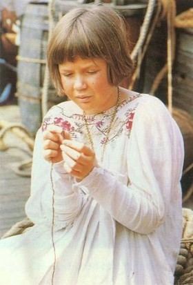 Sophie Wilcox is looking straight at the needle while trying to put the thread in it. Sophie with short blonde hair and bangs wearing a long necklace and long-sleeved medieval shift dress