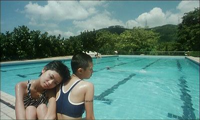 In the movie scene of Spacked Out 2000, in outdoor athletic size pool with trees at the back, from left, Wing Cheung is serious, sitting on at the side of the pool, leaning to Maggie Poon’s back, has black short hair, wearing a animal printed black and white swimsuit, at the right, Maggie poon is serious, sitting beside the pool, has black bald hair wearing, a black swimming bra.