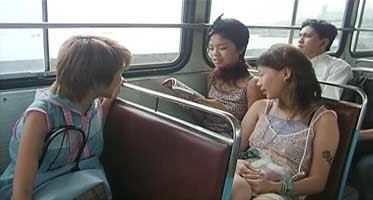In the movie scene of Spacked Out 2000, inside a public bus with stainless seat’s with maroon back, from left, Debbie Tam is serious, sitting with a blue shoulder bag at her lap, looking at her back, has brown short hair wearing a blue with pink shirt, 2nd from left, Wing Cheung is smiling, sitting while reading a book with her hands, has short black hair, wearing a red fur scarf on her neck and a printed noodle strap top, 3rd form left, Angela Au is serious, sitting, looking a the book, hands together on her lap, has short brown hair wearing a printed noodle strap top. At the right, a man is serious, sitting, looking outside the window, has black hair wearing a white shirt.