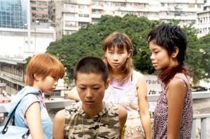 In the movie scene of Spacked Out 2000, From left, Debbie Tam is serious, standing in a white foot bridge, with a blue shoulder bag on her right, has brown hair wearing a blue shirt, 2nd from left, Maggie Poon is serious, looking down, standing, has black bald hair wearing a camouflage shirt, 3rd from left, Angela Au is serious, standing hands down, has brown hair, wearing a white dress, At the right, Christy Cheung is serious, standing, looking at Maggie Poon(2nd), has short black hair with a hair clip, wearing a printed shirt with a brown fur scarf on her neck.