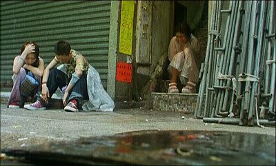 In the movie scene of Spacked Out 2000,  from left, Debbie Tam is sad, crouching with a blue shoulder bag, right hand on her head, and left hand under her chin, has brown hair wearing a bracelet, black shirt and gray pants with red shoes, 2nd from left, Maggie Poon is serious, sitting on the ground in front of a store with roll up cover, looking down, has black bald hair, wearing a camouflage shirt, black bracelet and a denim pants with red shoes, at the right, Angela Au is crying, sitting on the stairs, looking down, has black hair, wearing a white shirt, white pants and white shoes.