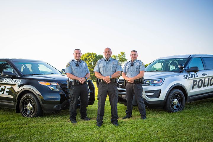 Has clear blue sky in a grassland with trees at behind two SUV’s from left, a black police SUV with police markings “Sparta” at the right is a silver police SUV with police markings “Sparta” in front is three police men, from left a man is smiling, standing with his hand together in his belt, has black hair, wearing a blue Sparta police uniform with a black radio on his right chest, black watch and a black pants with a gun on his right belt along with black shoes, holster, in the middle a man is smiling, standing with his hand together in his belt, has bald head, white beard, wearing a blue Sparta police uniform with a black radio on his right chest, silver watch and a black pants with a gun on his right belt holster, along with black shoes, at the right a man is smiling, standing with his hand together in his belt, has black hair, wearing a blue Sparta police uniform with a black radio on his right chest and a black pants with a gun on his right belt holster, along with black shoes,