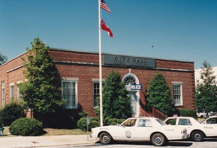 The City Hall of Sparta, Mississippi, the top Has clear blue sky, in the middle a building with red brick wall, has white windows, a white door with a blue police sign and large pine trees and round bushes, in front is a flag pole with a red flag under a USA flag, a parking lot with three white police box type car with police detailed marks on it.