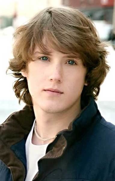 Spencer Treat Clark with curly hair while wearing a brown and blue vest, white t-shirt, and necklace