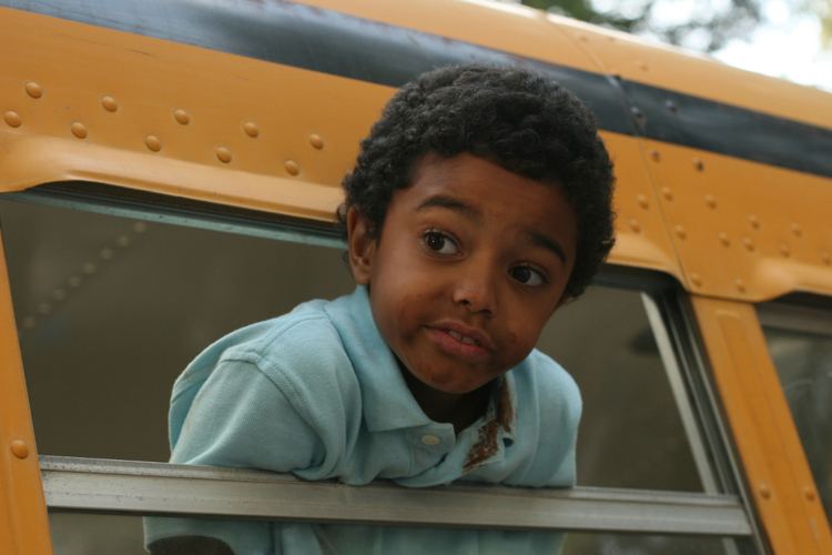 Spencir Bridges looking afar while leaning outside the window of a yellow bus in a scene from the 2007 American comedy film, Daddy Day Camp having curly hair and a smudge on his face and dirt on his collar, wearing a baby blue collared shirt.