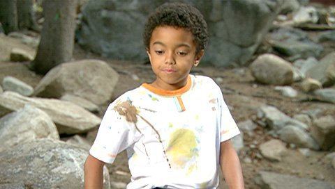 Spencir Bridges looking serious looking at the ground with rocks in the background in a scene from the 2007 American comedy film, Daddy Day Camp having black curly hair, wearing a stained white shirt.