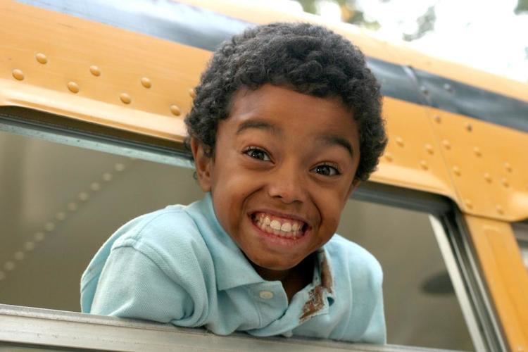 Spencir Bridges smiling showing his teeth while leaning outside the window of a yellow bus in a scene from the 2007 American comedy film, Daddy Day Camp having curly hair and a smudge on his face and dirt on his collar, wearing a baby blue collared shirt.