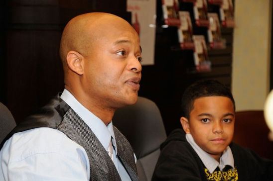 Spencir Bridges looking serious while sitting next to his father, Todd Bridges talking with someone, and bookshelf in the background during Todd's new book signing "Killing Willis" on March 23, 2010 in Los Angeles, California. Spencir is wearing a black shirt with prints on under a black hoodie jacket while Todd is wearing a white satin necktie, a light-blue collared shirt under a patterned dark-gray vest.