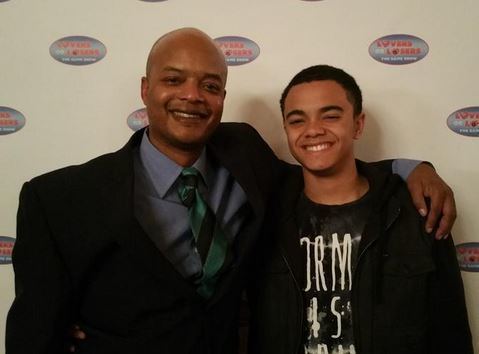 Spencir Bridges with his father, Todd Bridges both smiling while Todd is wrapping his arm on Spencir's shoulder with a banner in the background. Todd has a bald head, wearing a green and black striped necktie, a blue collared long sleeve under a black coat, while Spencir has black clean-cut curly hair, wearing a printed black shirt under a black zipper jacket.
