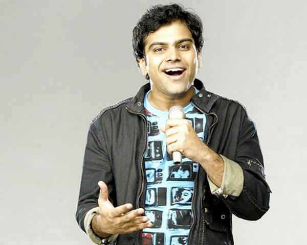 Sreerama Chandra Mynampati smiling and holding a microphone while wearing a black jacket over a sky blue shirt.