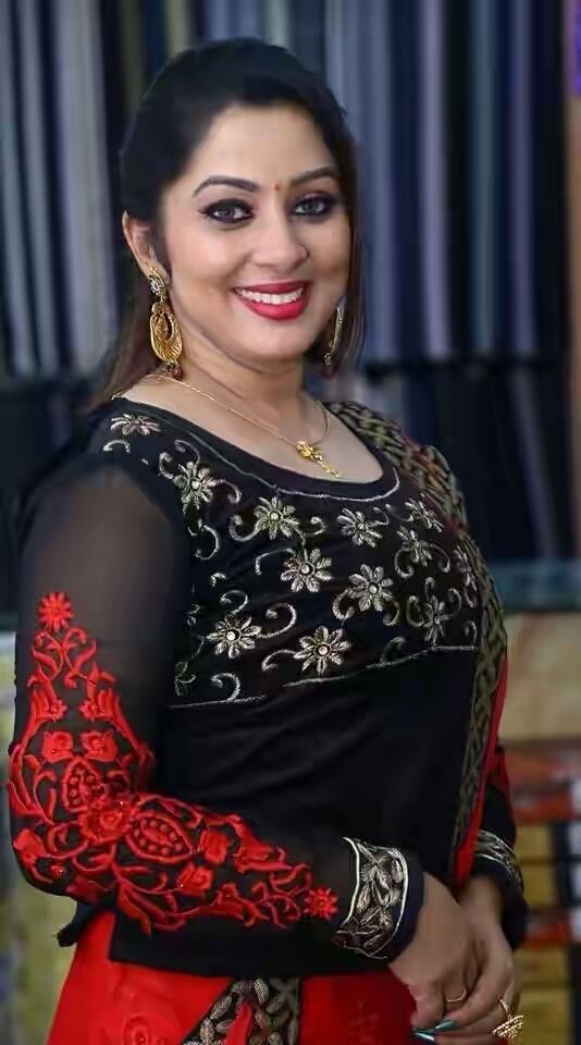 Sreeya Remesh smiling with a bindi on her forehead, she has a black hair wearing dangling earrings, a gold necklace, a ring on her right ring finger so as her left middle finger, and black and red dress
