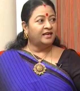 Sri Lakshmi speaking and wearing a black and violet saree with a red mark on her forehead, a gold necklace and dangling earrings