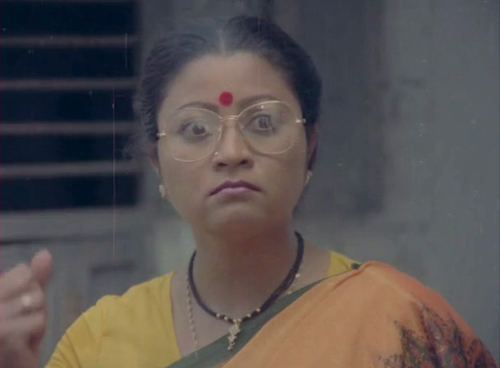 Sri Lakshmi showing a serious face with a red mark on her forehead while wearing a yellow saree, eyeglasses and necklace