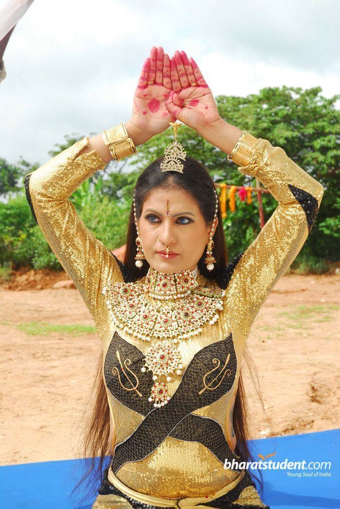 Chandrika looking at something and raising her hands, with a serious face and red marks on her hand, while wearing a black and gold long sleeve full bodysuit, small gold crown, plastron necklace, nose-jewel, bracelet, and earrings