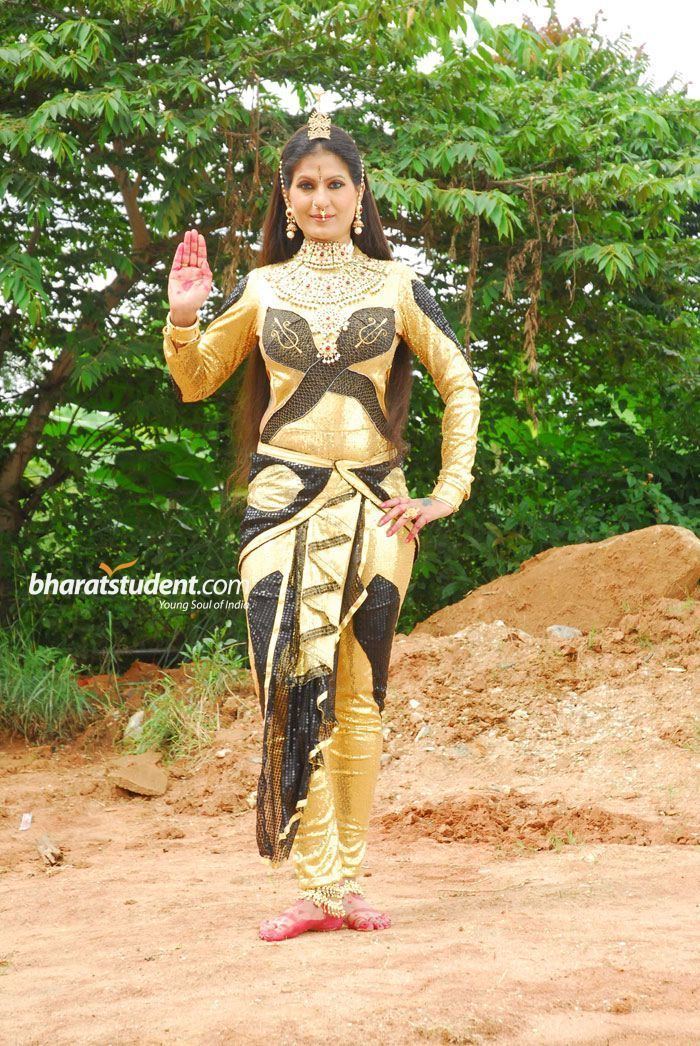 Chandrika with a tight-lipped smile while raising her left hand and right hand on her waist, with red marks on her hands and feet. She is wearing a black and gold long sleeve full bodysuit, a black and gold cloth wrapped around her waist, a small gold crown, ankle bracelet, plastron necklace, nose jewel, bracelet, and earrings