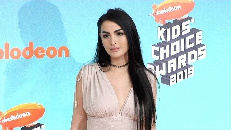 SSSniperWolf with a serious face, long black hair, wearing a black choker, and a beige sleeveless top.