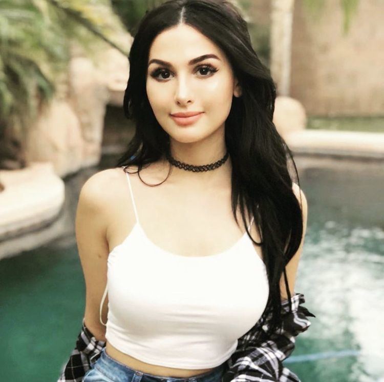 SSSniperWolf smiling, with long wavy black hair, wearing a black choker, a white spaghetti top with a checkered black and white shirt, and blue pants.