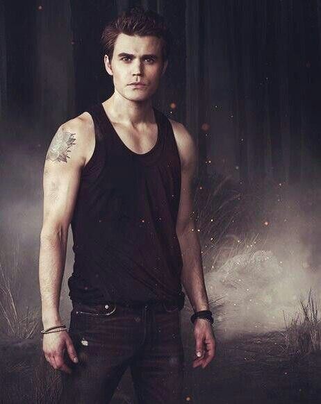 Stefan Salvatore with a tight-lipped smile, with a tatoo on shoulder and wearing a sliveless t shirt , brown spiked hairs while wearing a black banyan and black jeans