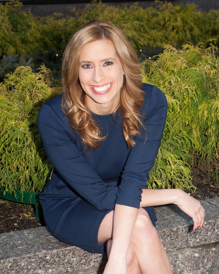 Stephanie Abrams smiling while sitting on a bench with crossed legs, wavy blonde hair, and wearing a dark-blue dress.