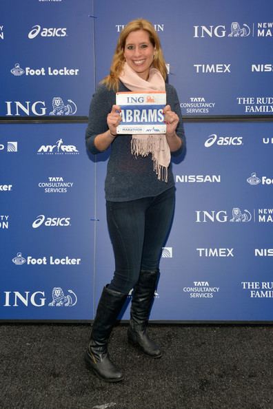 Stephanie Abrams smiling while holding a placard, with wavy blonde hair, wearing an off-white scarf, gray sweatshirt, dark blue pants, and black boots.