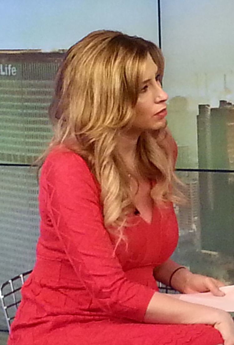 Stephanie Abrams' side-view while sitting on a chair, with long wavy blonde hair, and wearing a red-orange dress.