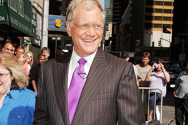 David Letterman is smiling while looking at someone, a late-night host of the Late Show with David Letterman. With a crowd and buildings in the background, he has white hair, eyeglasses, a violet polka dot necktie with a microphone, wearing a white collared shirt, and a black pinstriped suit.