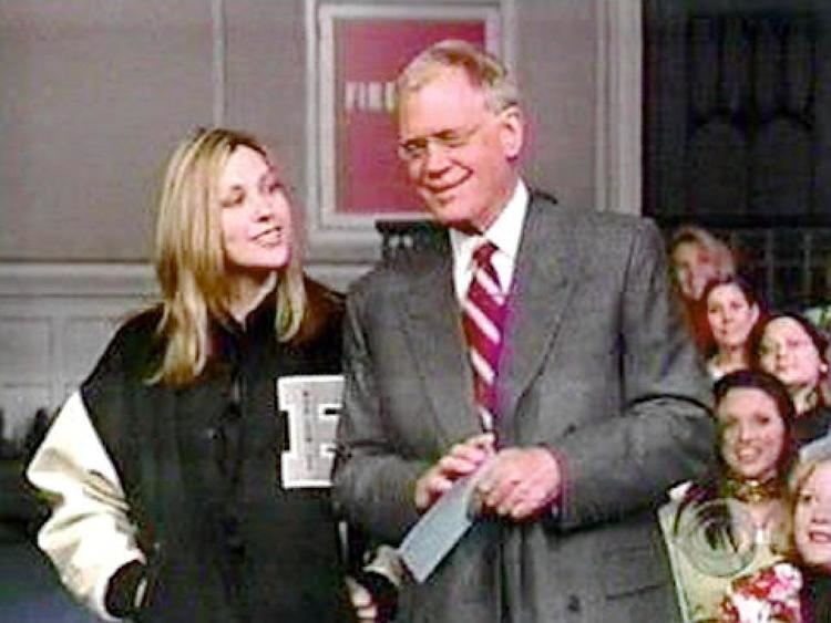 Stephanie Birkitt and David Letterman smiling while David is holding a letter and Stephanie have her hands in her pocket with people in the background. Stephanie has wavy blonde hair wearing a black and white varsity jacket while David with blonde hair is wearing eyeglasses, a red and white stripes necktie, a white collared shirt, and a gray coat.