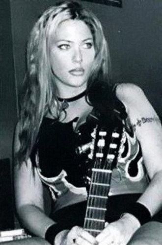 Stephanie Opal Weinstein with a serious face while holding a guitar, with long blonde hair, wearing a choker, and a black sleeveless top.