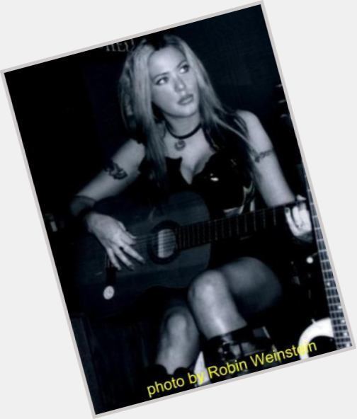 Stephanie Opal Weinstein with a serious face while playing the guitar, with blonde hair, with tattoos on her arms, wearing a choker, a black sleeveless sexy dress, and boots.