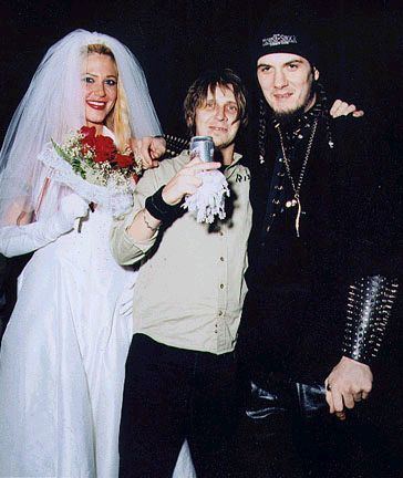 Stephanie Opal Weinstein smiling, wearing a wedding dress while holding a bouquet of red roses, and Phil Anselmo also smiling, wearing a necklace, a black hat, a black shirt, and black pants with his arm around the shoulder of a man holding a can of beer and wearing a long sleeve and black pants.