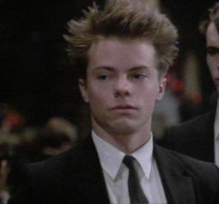 Stephen Geoffreys wearing a black coat, white long sleeves, and black necktie in a scene from the 1985 film, Heaven Help Us