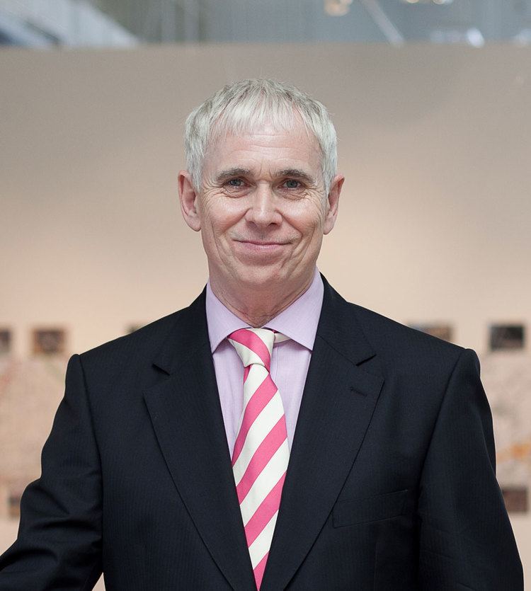 Steve Dixon smiling closed mouth and wearing a black coat over a light pink suit and a striped necktie.