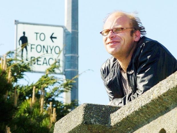 Steve Fonyo smiling while looking afar with signage on the left side saying "To Fonyo Beach". Steve is wearing eyeglasses and a brown shirt under a black leather jacket