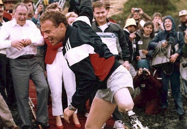 Steve Fonyo smiling while running with a prosthetic leg and people are laughing at him. Steve is wearing a black, white, and red jacket and gray shorts