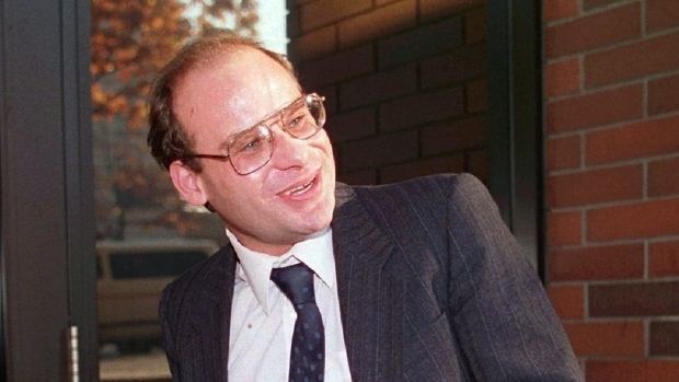 Steve Fonyo smiling while looking at something while wearing eyeglasses and a white long sleeve under a blue necktie and black striped coat