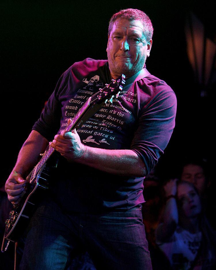 Steve Jones is serious, has black and white hair, a beard and mustache, both hands holding a guitar, with boys and girls at the back, he is wearing a printed black long-sleeve shirt, and black pants.