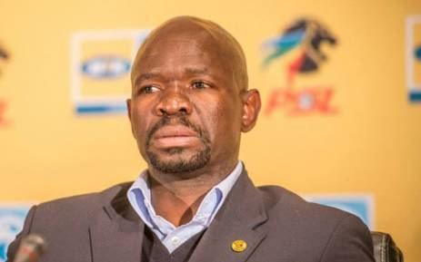 Steve Komphela sitting on a black couch with a serious face, with a microphone in front of him, wearing a gray coat with a golden badge over a gray vest, and light blue long sleeves.