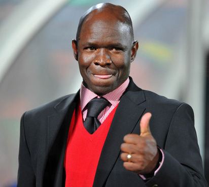 Steve Komphela is licking his lip while doing thumbs up, with a naughty smile, wearing a ring, a black coat over a red vest, striped pink long sleeves, and a black necktie.