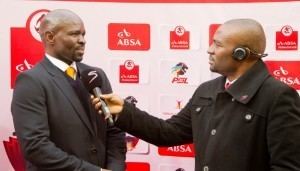 Steve Komphela is talking during an interview with a reporter holding a microphone. Steve with beard and mustache, wearing a gray coat over a black vest, white long sleeves, and a yellow tie while the reporter is wearing a wireless microphone on his head and a black coat over white long sleeves.