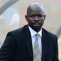 Steve Komphela with a serious face, with beard and mustache, wearing a black coat over white long sleeves, and a checkered light brown tie.