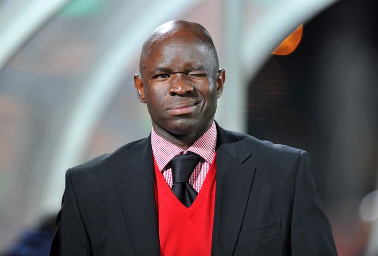 Steve Komphela is winking, wearing a black coat over a red vest, striped pink long sleeves, and a black tie.