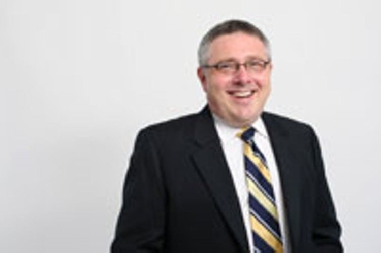 Steve Murphy with slight gray hair smiling on set and wearing a black coat over a white long-sleeve and yellow and blue necktie.