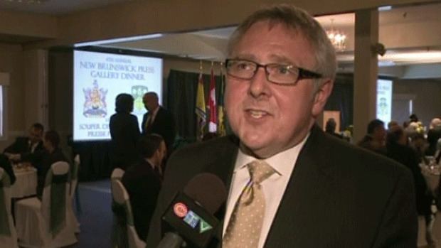 Steve Murphy speaking in an interview and wearing a white long-sleeved white suit and cream necktie underneath a black coat.