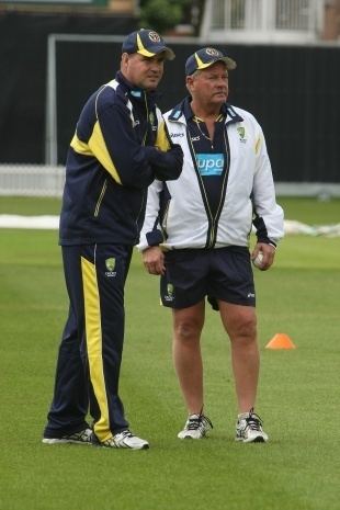 Steve Rixon (right) standing along with Jake White (left) inside of the field. Steve seriously looking from afar and holding a ball on his left hand, he has a white hair wearing a black cap, white shoes and a black polo jersey under a white jacket and black short while Jake seriously looking from afar with crossed arms wearing a black cap, white shoes, and a black polo jacket and long black pants